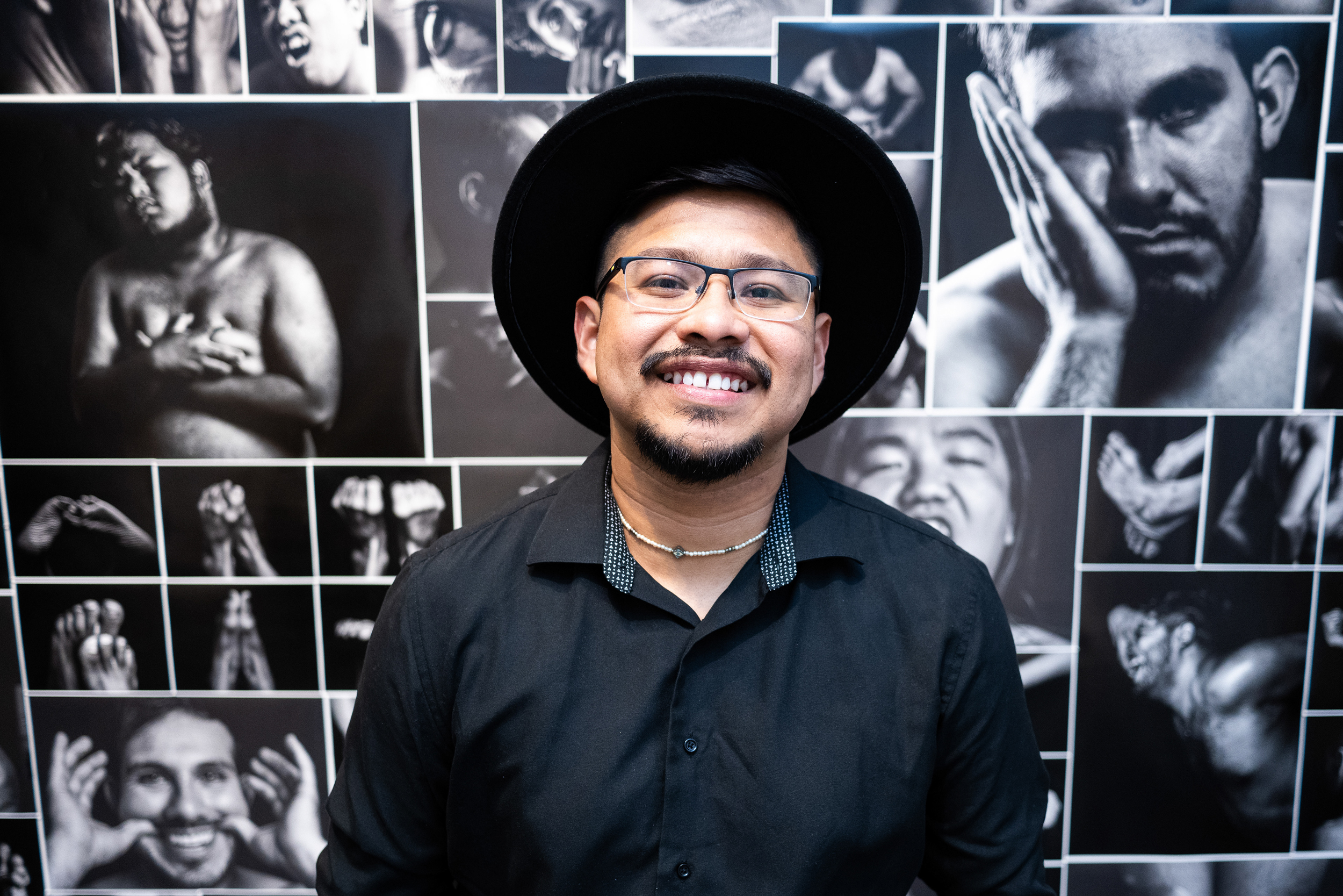 A male student with a goatee and glasses and a wide brimmed hat in front of a collage of black and white portraits