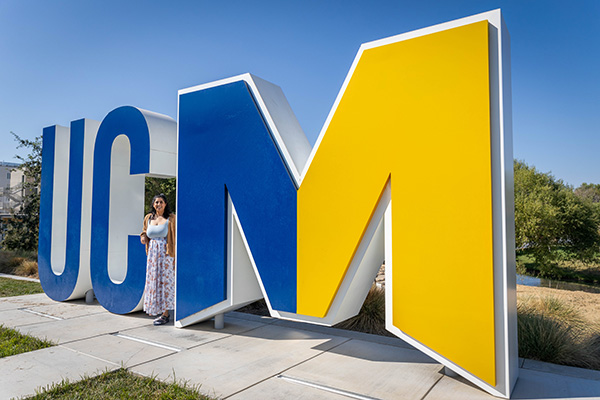 Woman in dress standing between the UC and the M in the UC Merced logo statue on campus