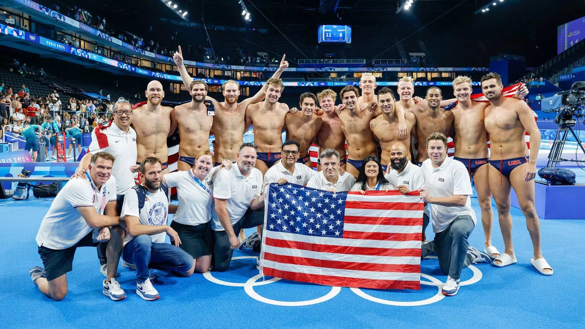 A group shot of the U.S. men's water polo team and staff, holding an American flag in front, celebrating their bronze medal in the aquatics center