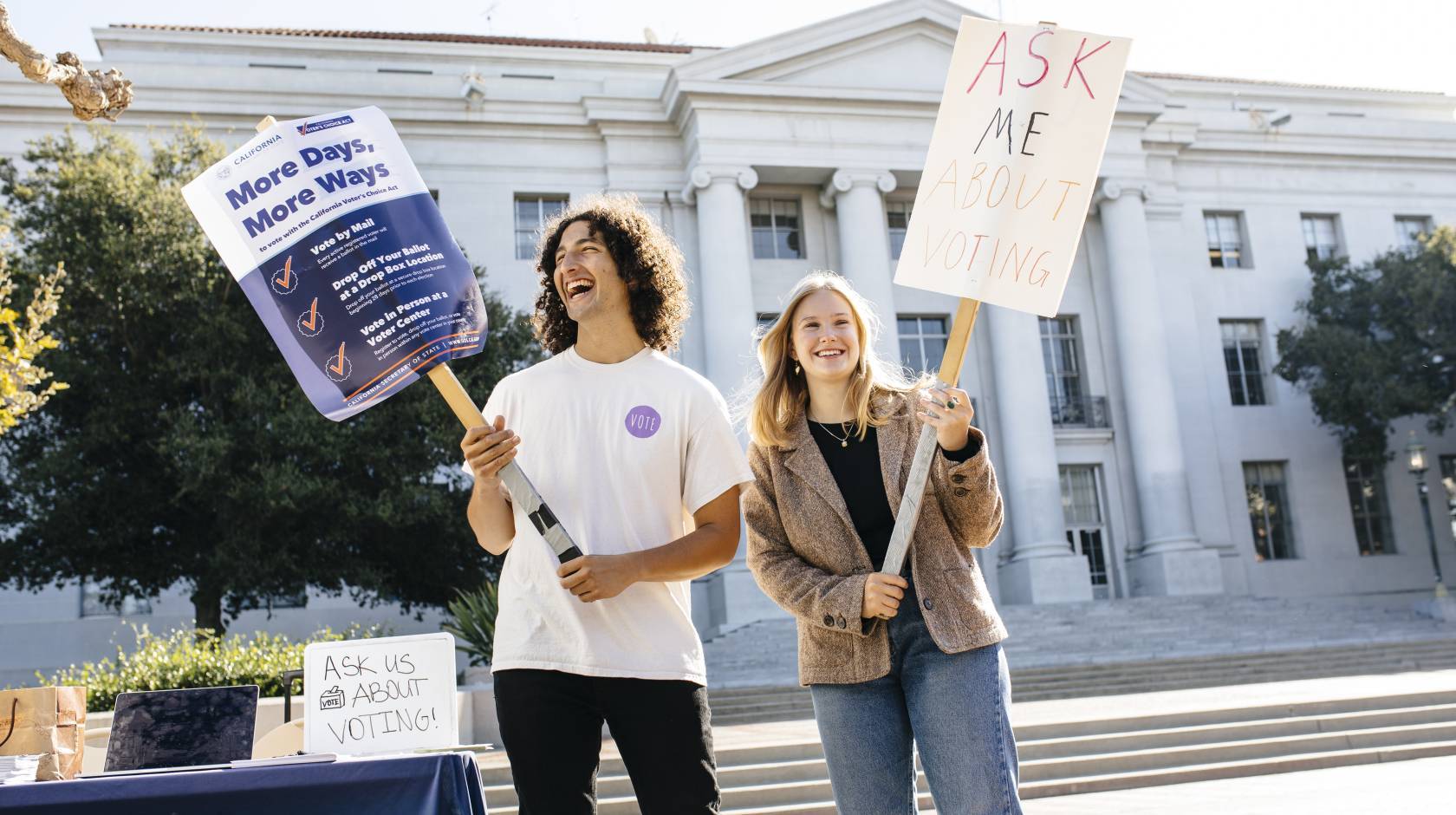 Two students hold signs on sticks with voting information displayed, standing in front of a white marble campus building