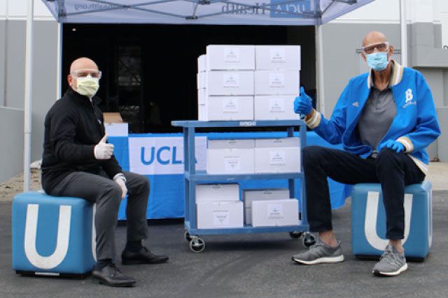 Kareem Abdul-Jabbar sits at a UCLA both wearing medical gloves, goggles and a surgical mask, giving a thumbs up