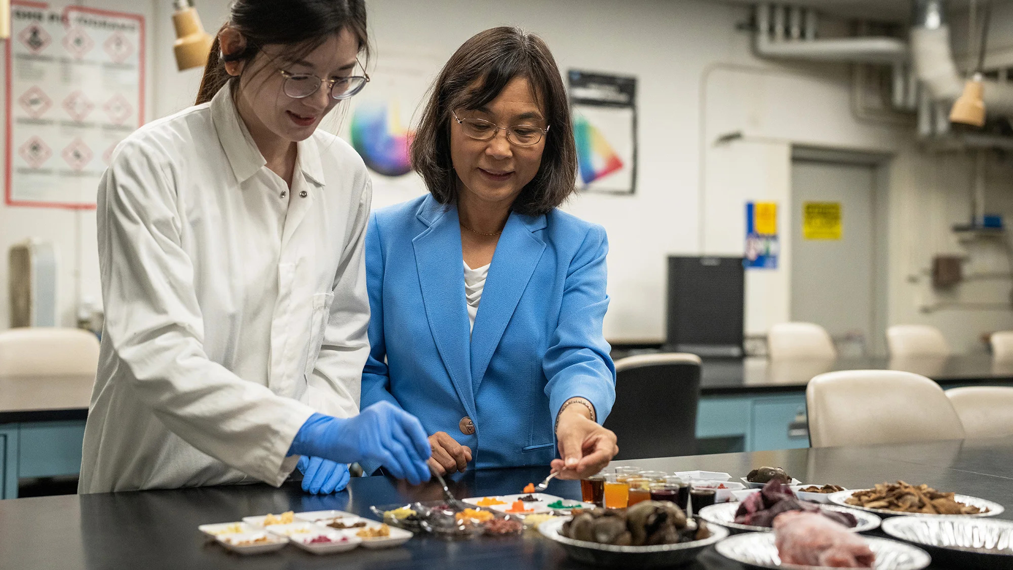 A woman researcher in a lab coat, left, and a woman in a sky blue blazer, right, lift up samples of small spherical colorful foods made of fungi in a lab