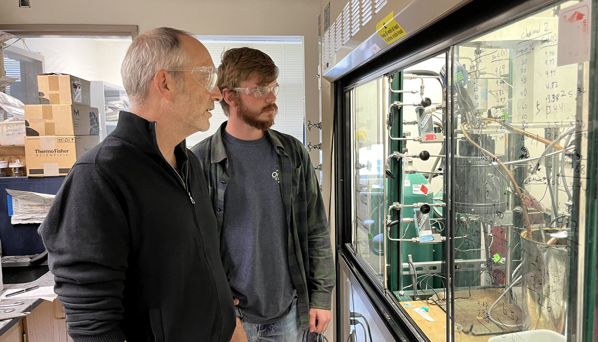 Two men look at a machine through a glass protective window