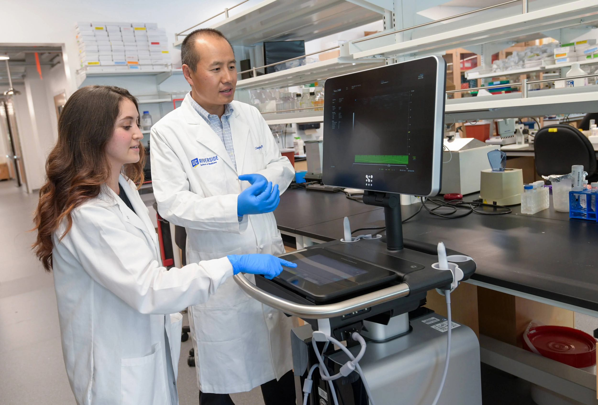 A woman in a lab coat with long hair and a man in a lab coat look at a screen -- both wearing blue medical gloves