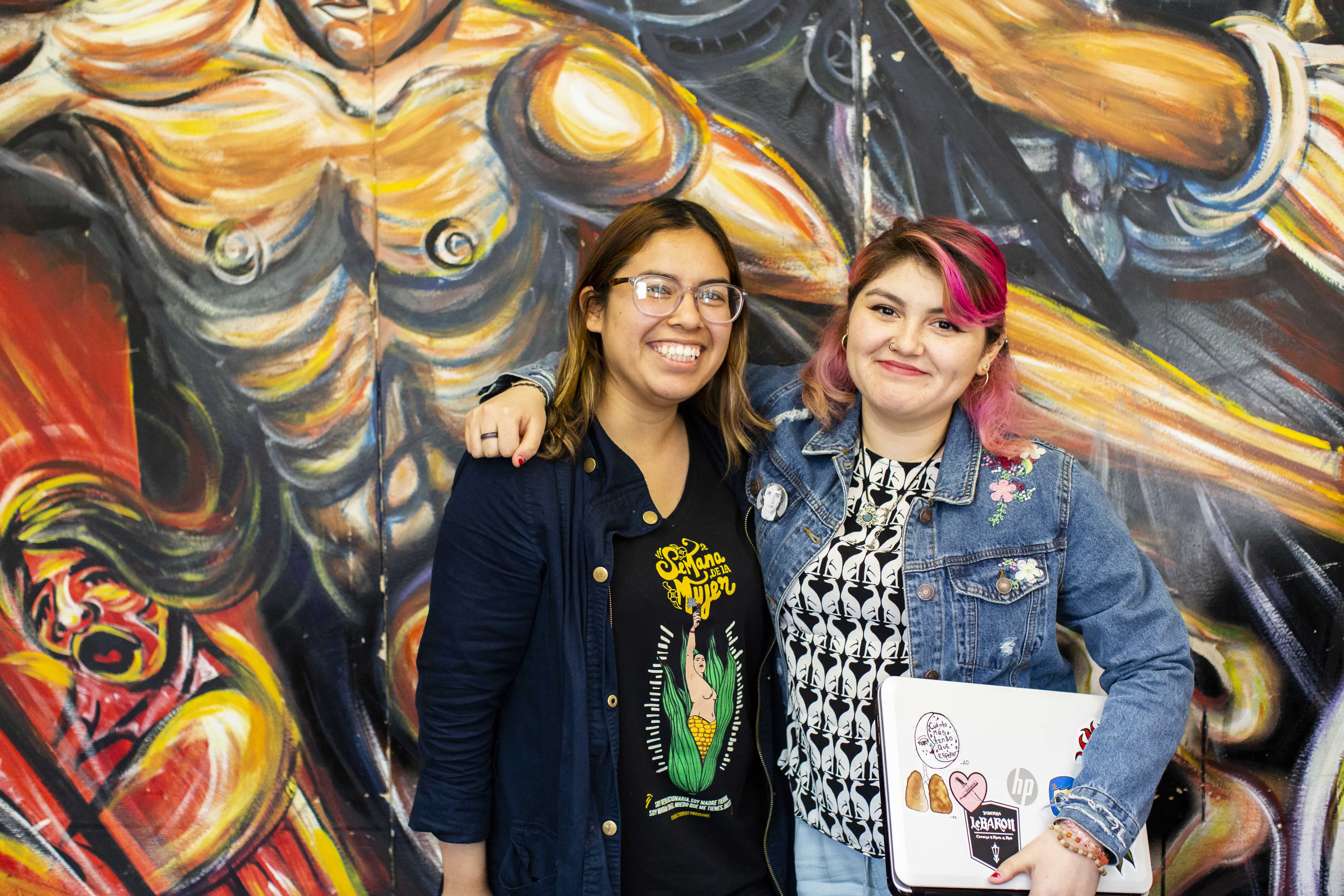 Two young women pose together in front of a colorful mural