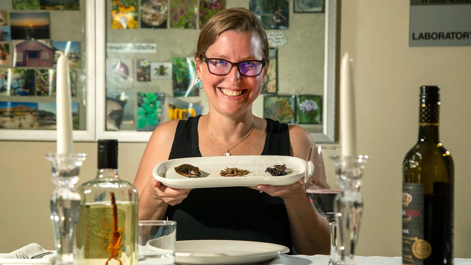 A grinning woman with glasses holds up a plate of bugs at a dinner table with olive oil and wine nearby