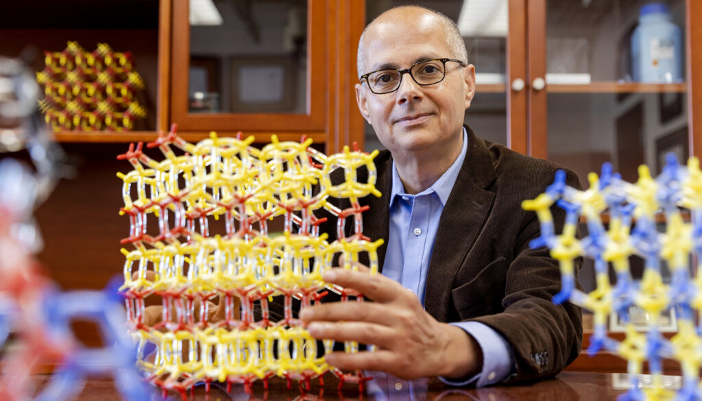 Man in tweed jacket in an office lined with wooden cabinets, holding a large model of a molecule assembled with smaller parts