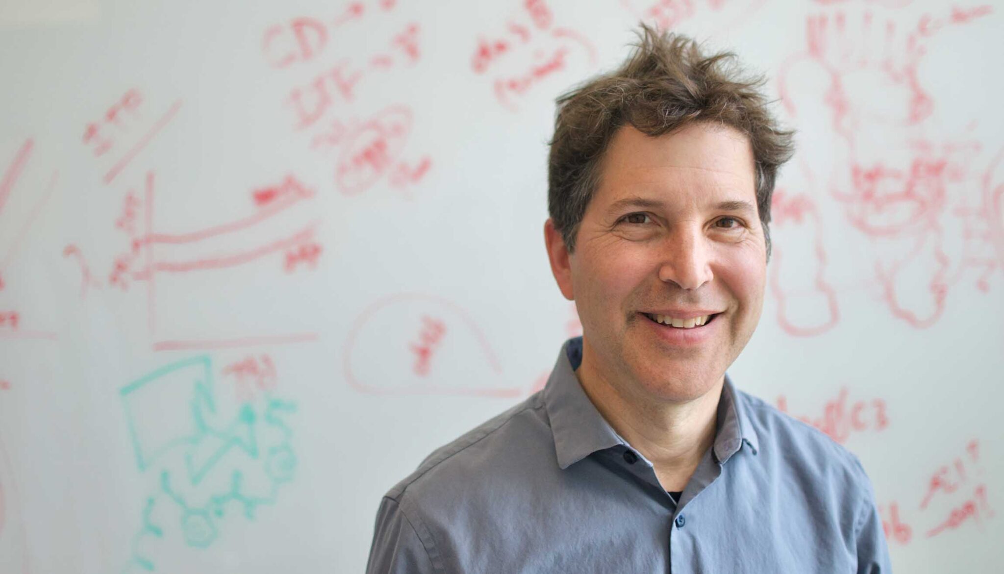 A man in a dress shirt smiles with a white board full of notes behind him