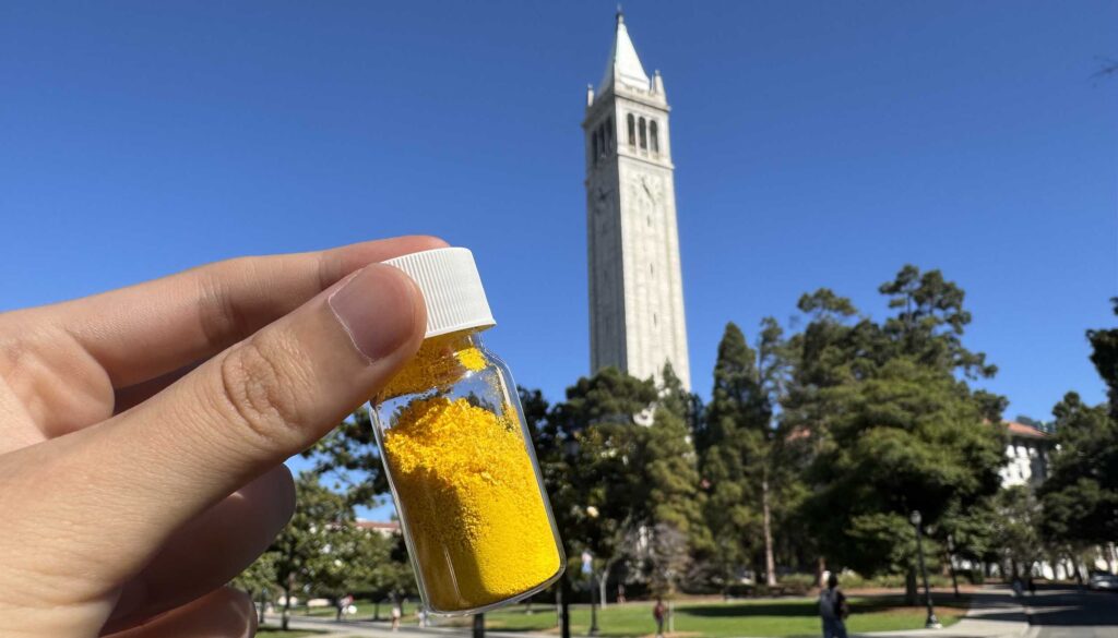 Hand holds a small vial of yellow powder up against a blue sky with Berkeley's belltower in the background