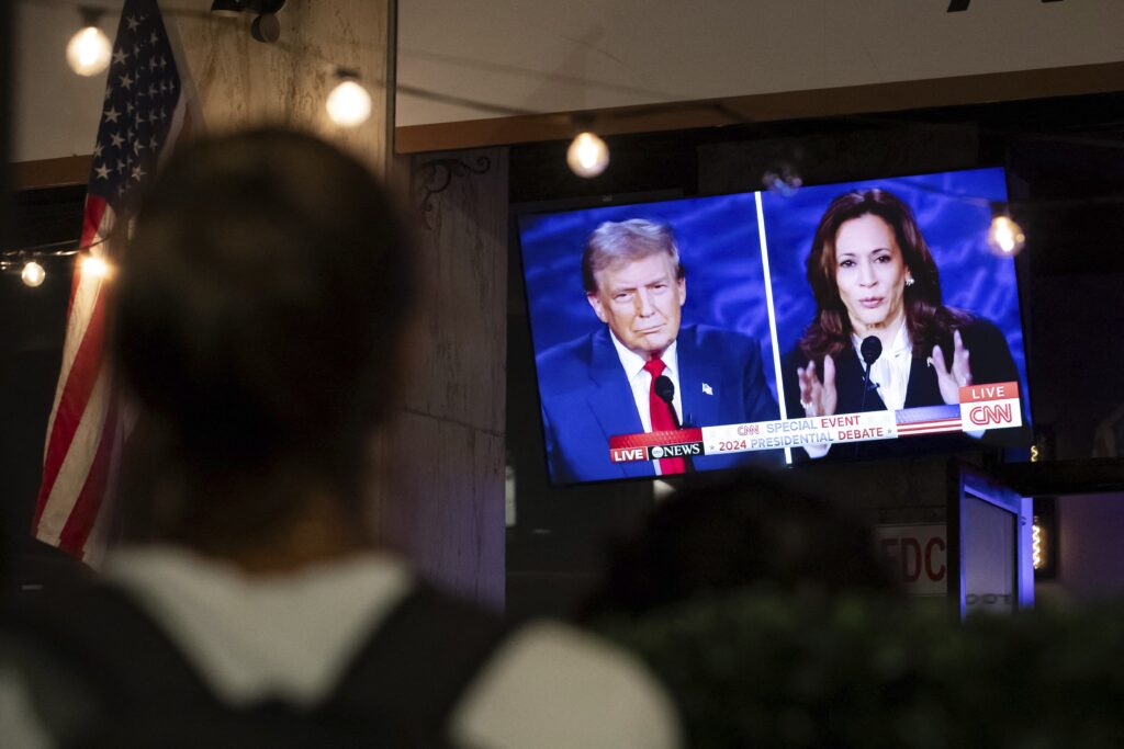 A split-screen image on a TV shows Donald Trump and Kamala Harris debating, being watched by people at a bar