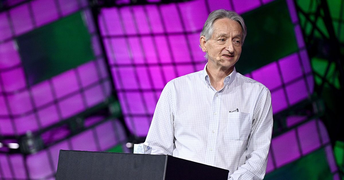 An older man gives a talk from a podium, a background of purple and green cubes behind him