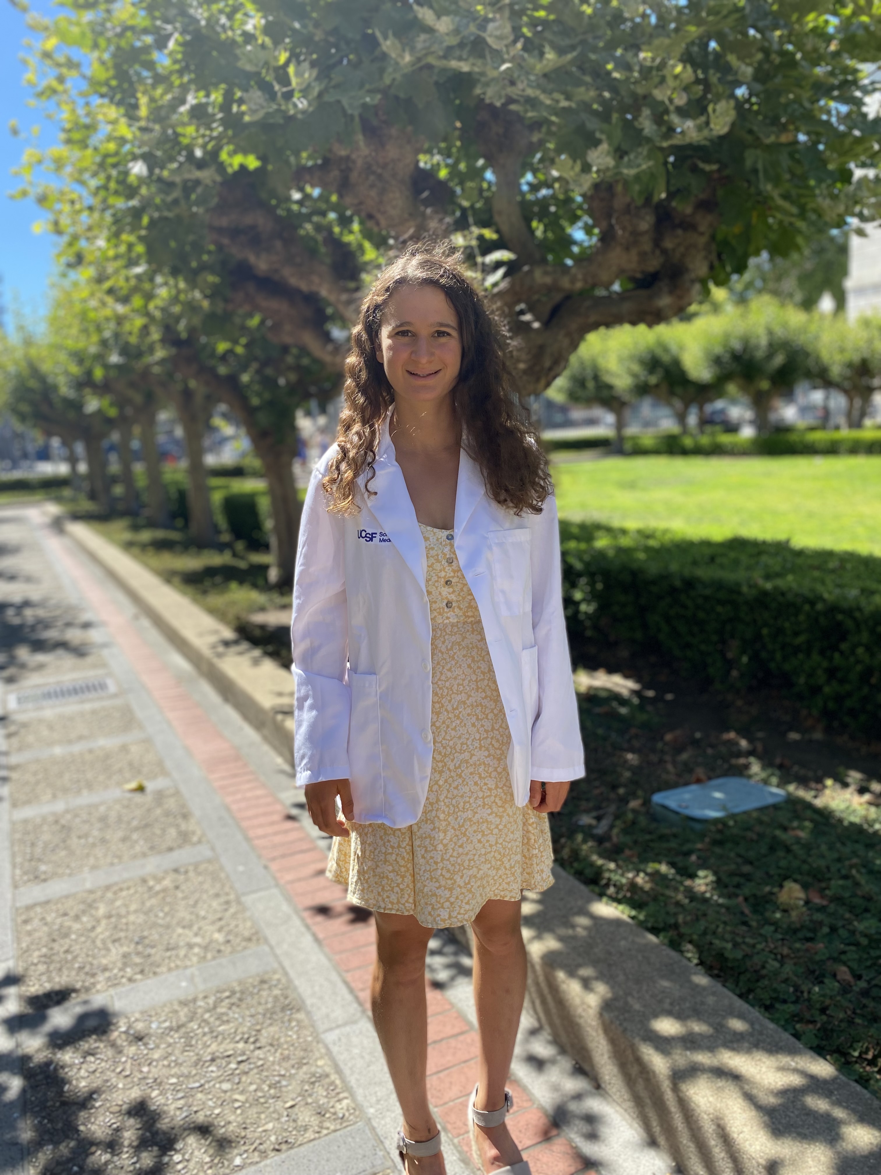 Talia Ruxin in a white physicians coat and dress, outside in front of trees and a lawn