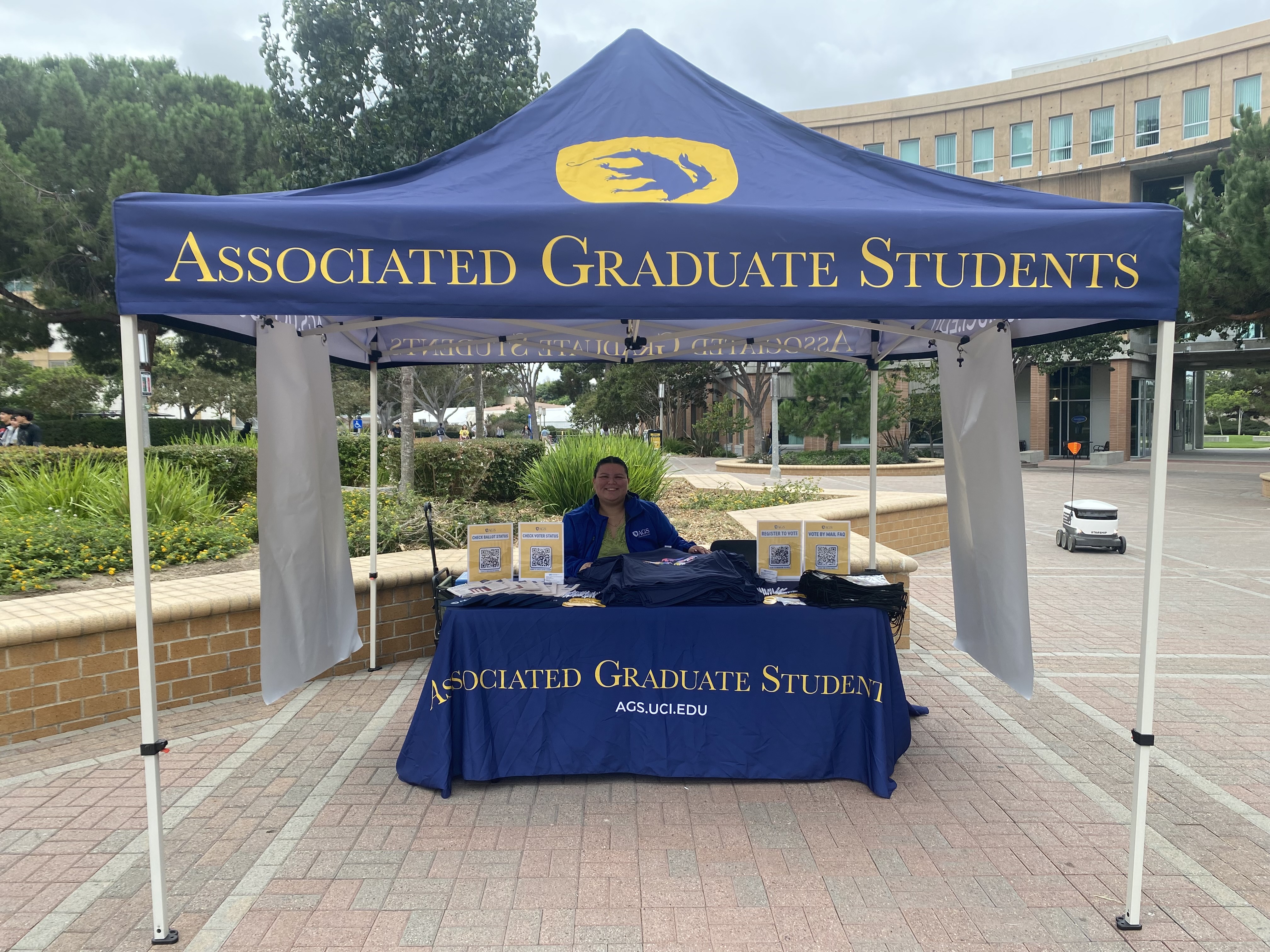 Rebecca Angela Ruiz sits at a table under a tent on campus with Associated Graduate Students branding