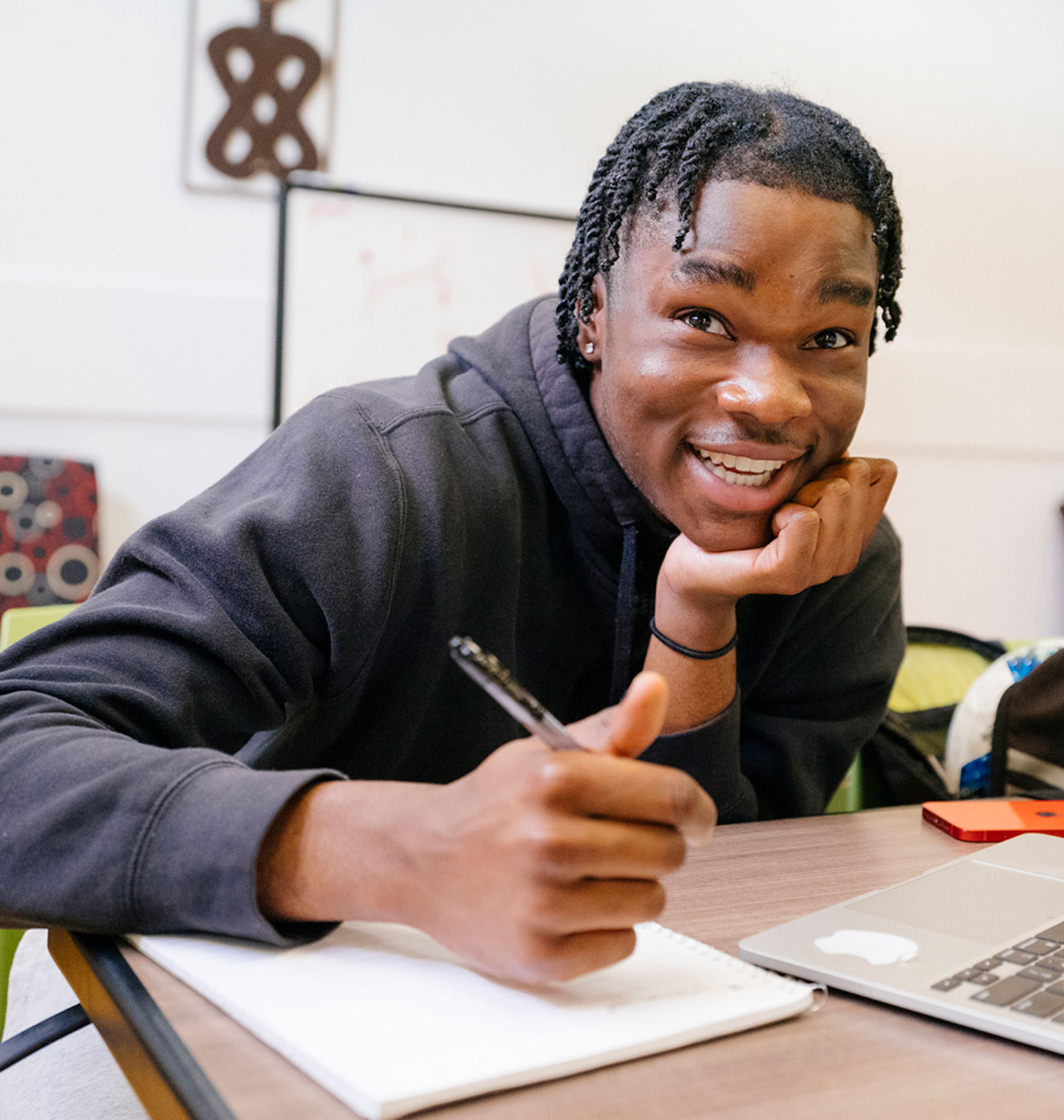 A college student sits in front of a laptop, chin propped on his hand, taking notes in a notebook with his other hand, looking up and the camera and smiling