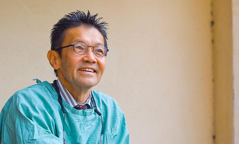 An older Asian man in a surgical gown and glasses smiles at something off in the distance