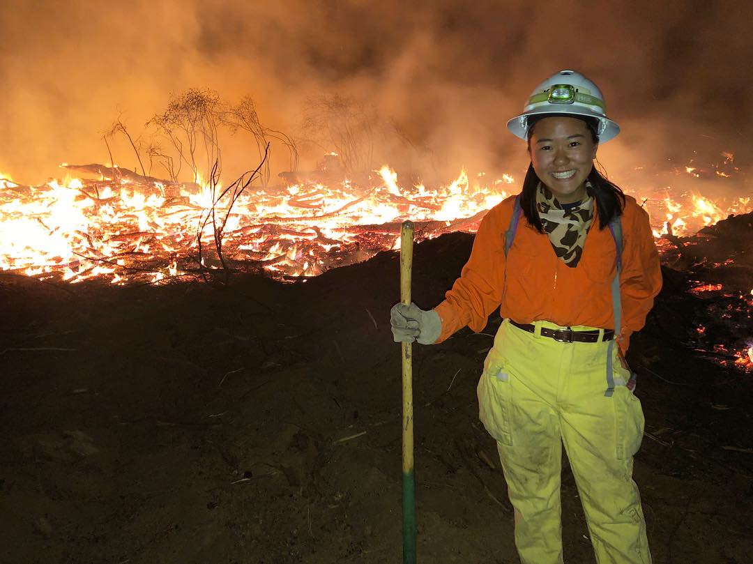 Katie Low wearing protective clothing and carrying a hand tool standing and smiling in front of a burning field 