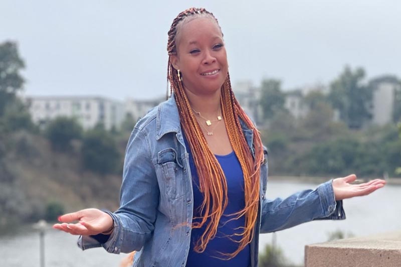 Woman with braids in a jean jacket holds her arms wide as if to show something on the UC Santa Barbara campus, lake in the background