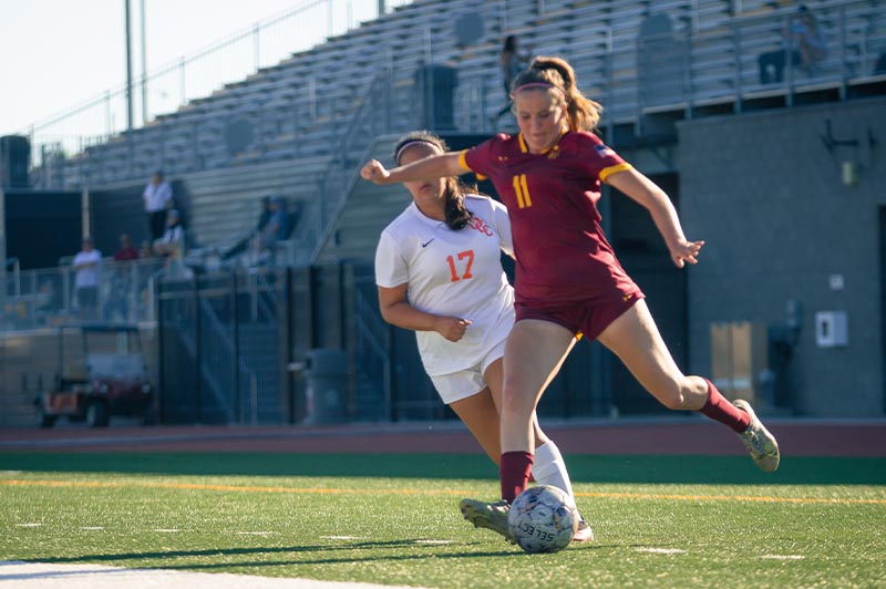 A young woman in a maroon jersey playing soccer gears up to pass the ball as a woman in a white jersey pursues her