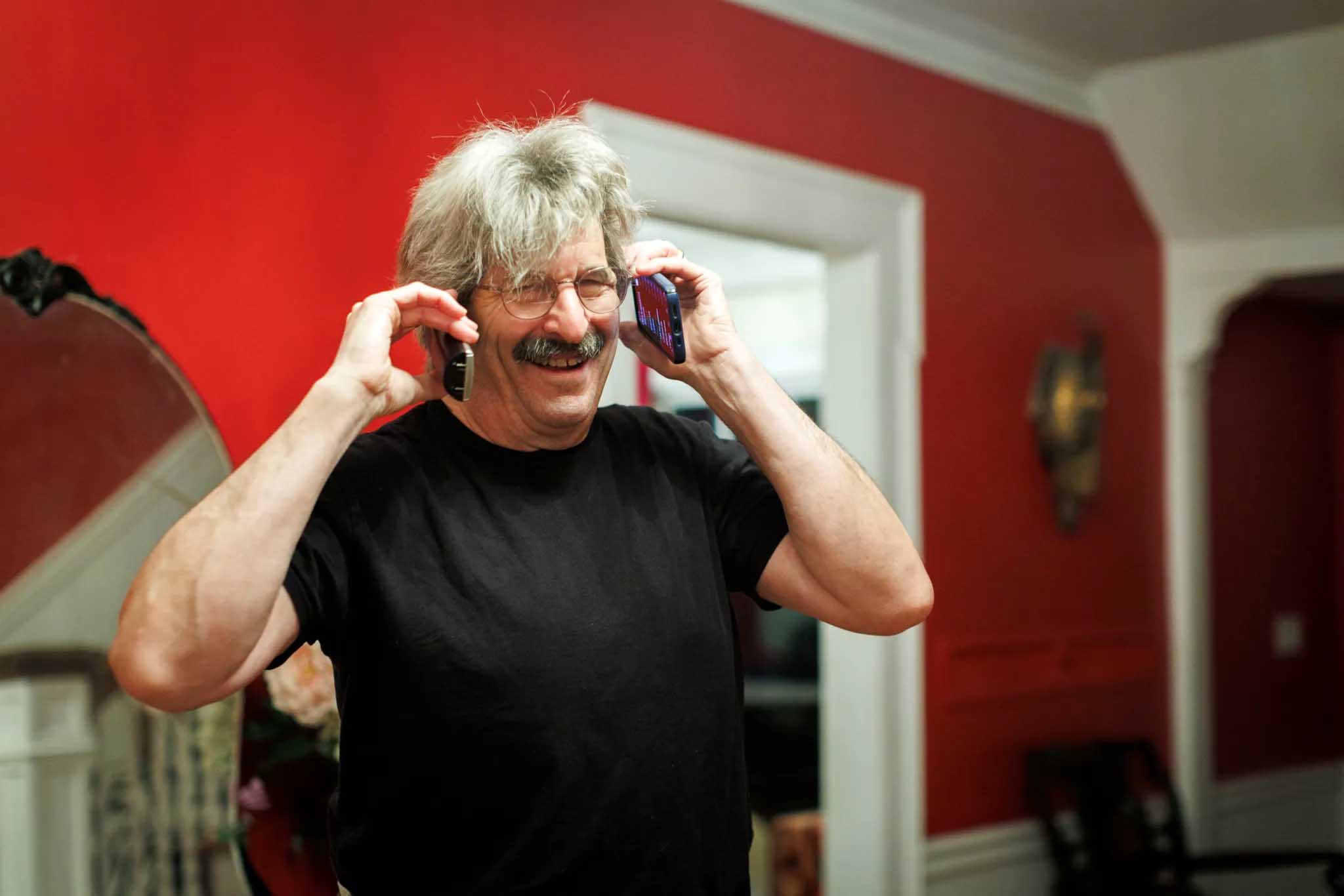 A man with bushy gray hair and a mustache smiles as he holds a phone to each ear in his house