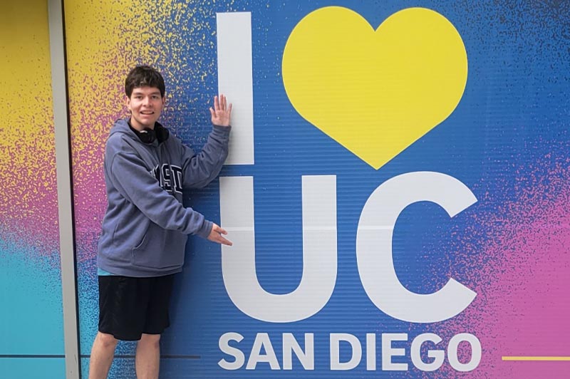 Young man in a hoodie and shorts next to an "I HEART UC San Diego" sign that he is gesturing toward while smiling