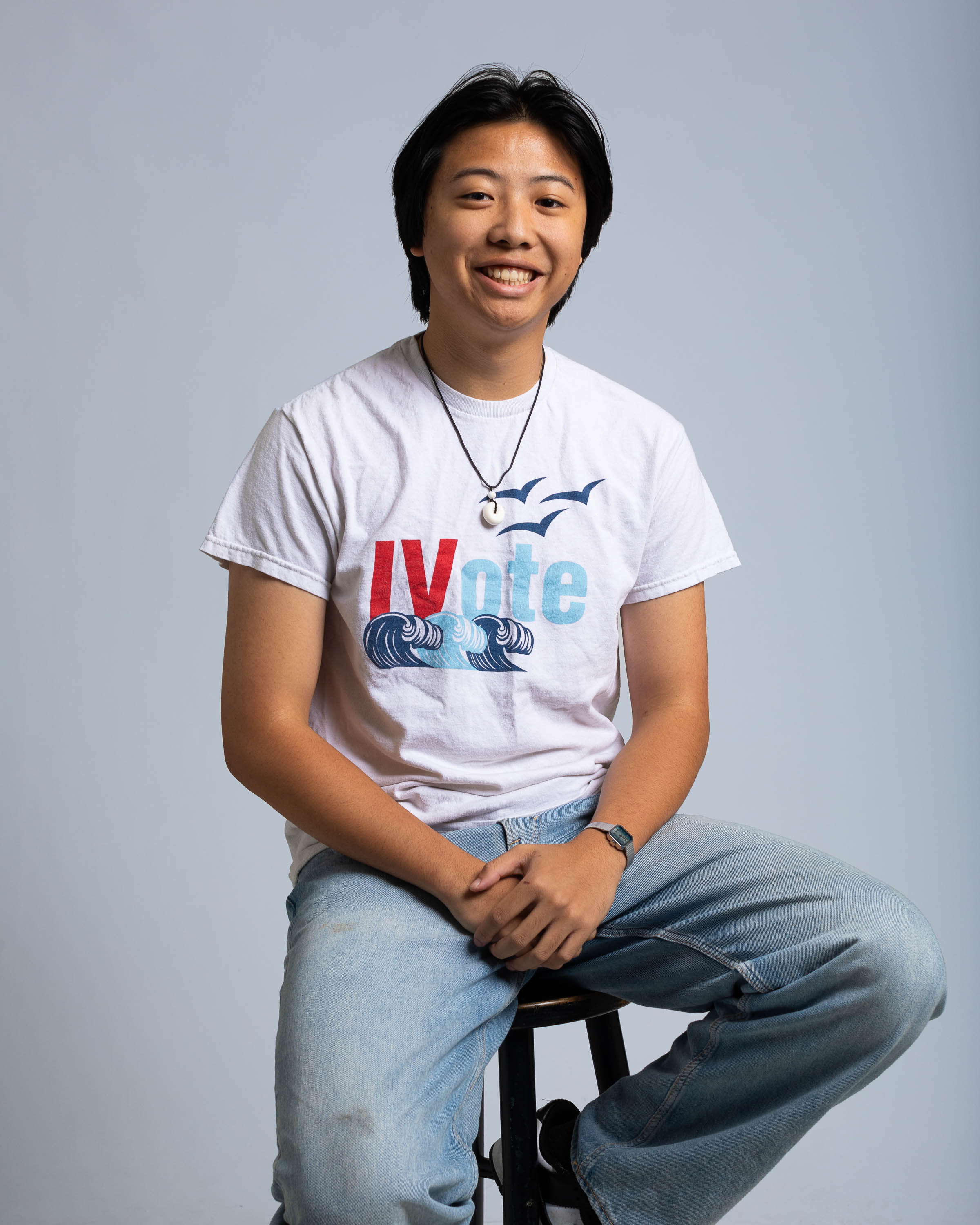 Byron Chan wearing a shirt reading "IV Vote" sits on a stool and smiles against a gray background