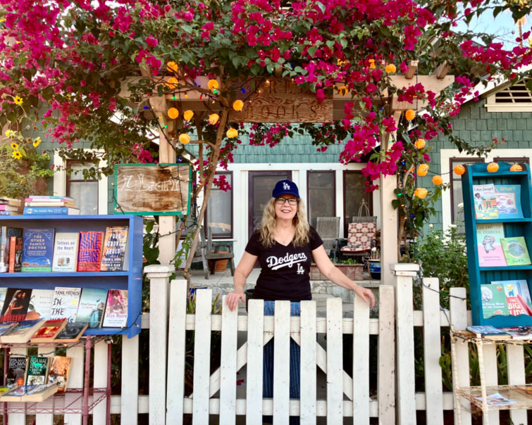 Susan Straight stands behind a white picket fence, between 2 Little Free Libraries, under a tree covered with bright pink blossoms