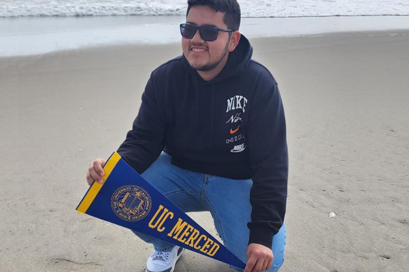 Young man in sunglasses holding a UC Merced pennant posing on one knee in front of the ocean