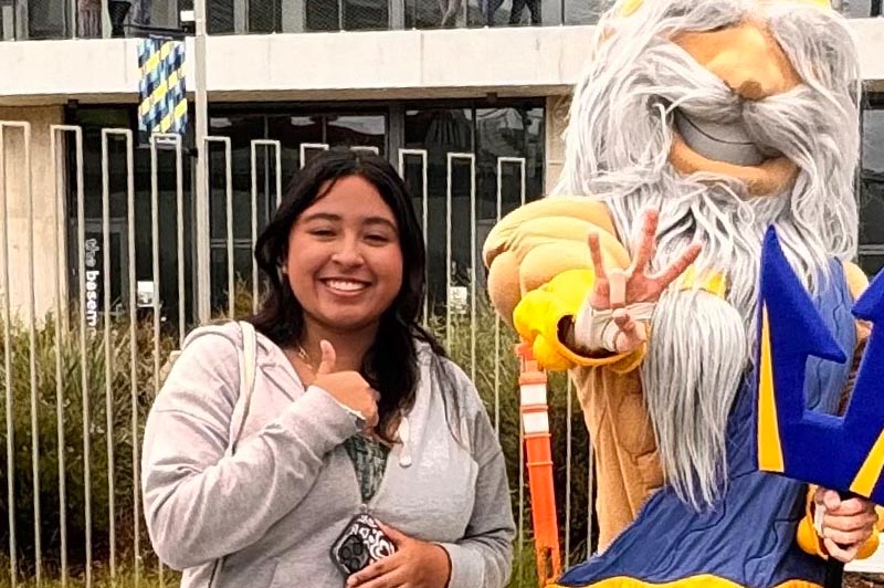 A young woman grins and gives a thumbs up sign next to the UC San Diego Triton mascot