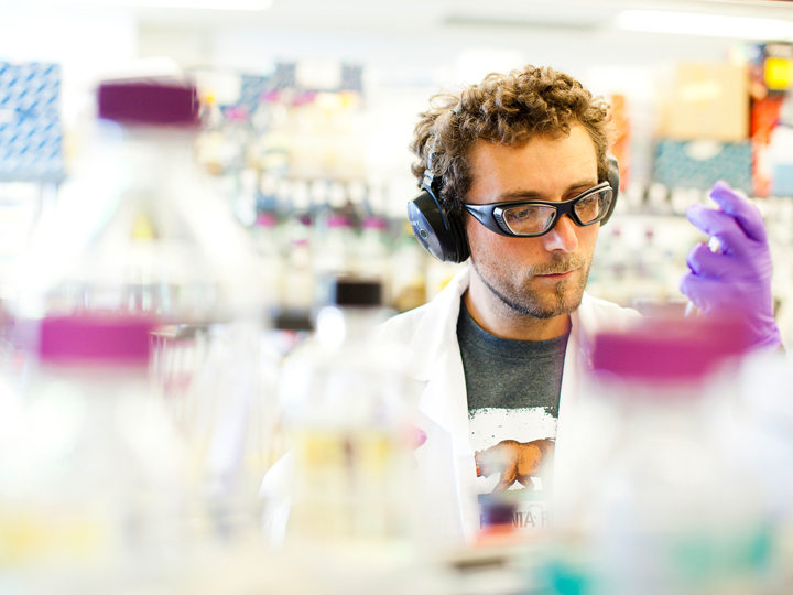 A young man pipettes in a lab wearing a California flag t-shirt, white lab coat, purple gloves, safety glasses and headphones