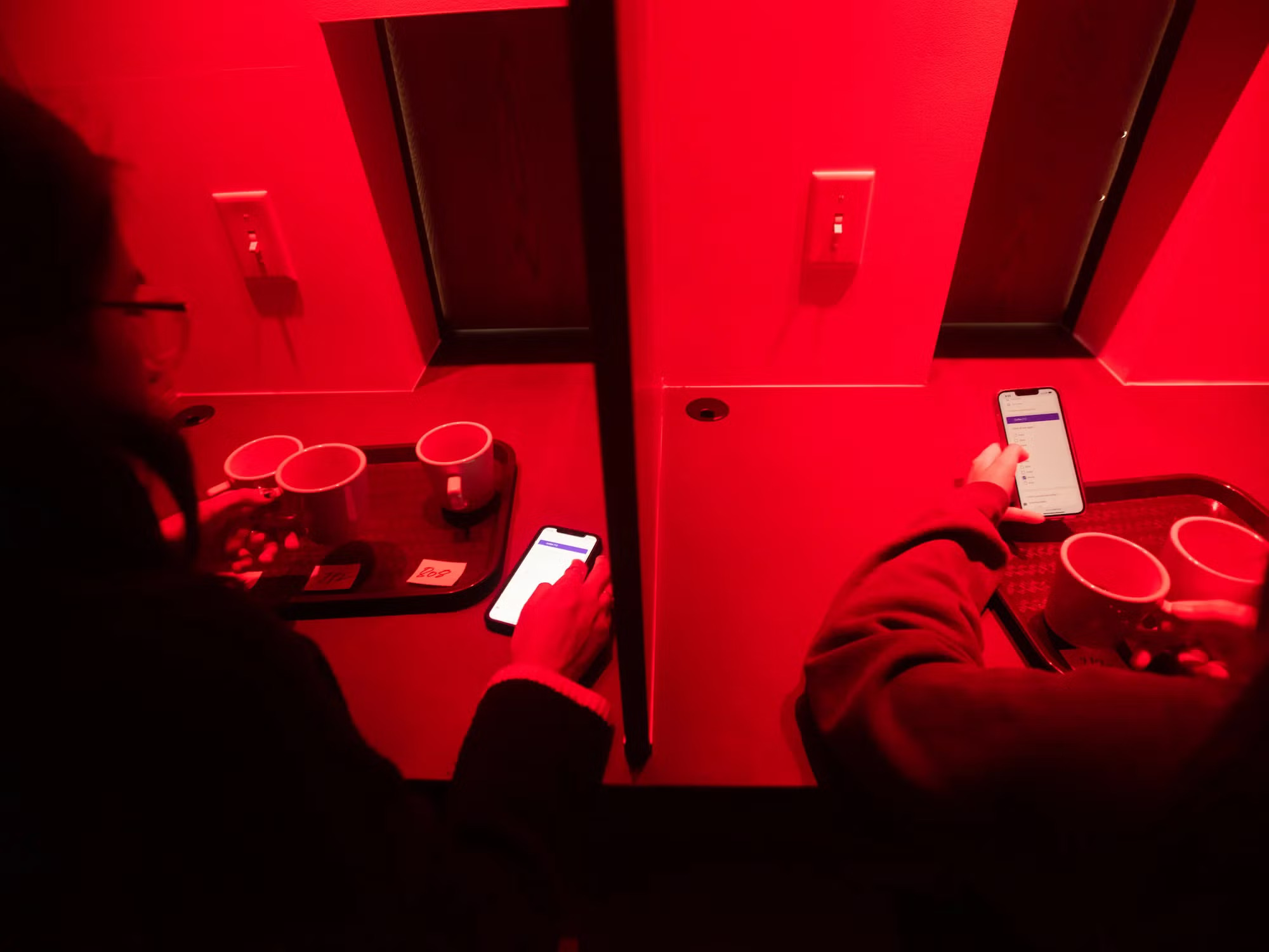 In two stalls with red lighting, two students (mostly not in frame) hold their phones and evaluate coffee cups in front of them