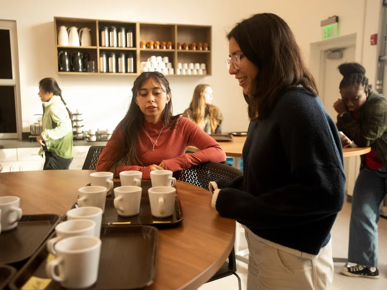 In a cafe with a few people in it, two young women look at a series of mugs on a tray