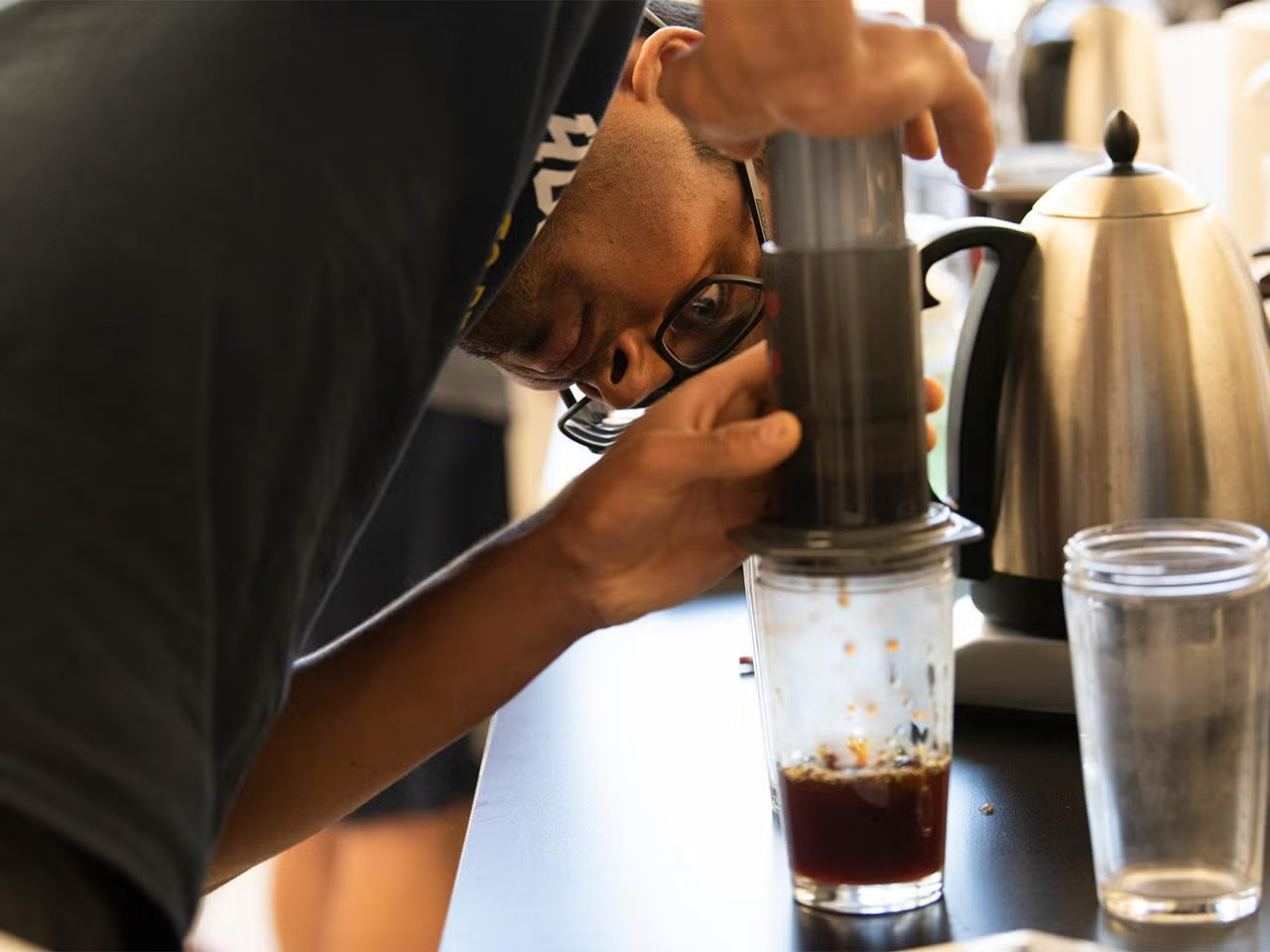 A young man with glasses pushes coffee through a tube into a glass, craning his head to look