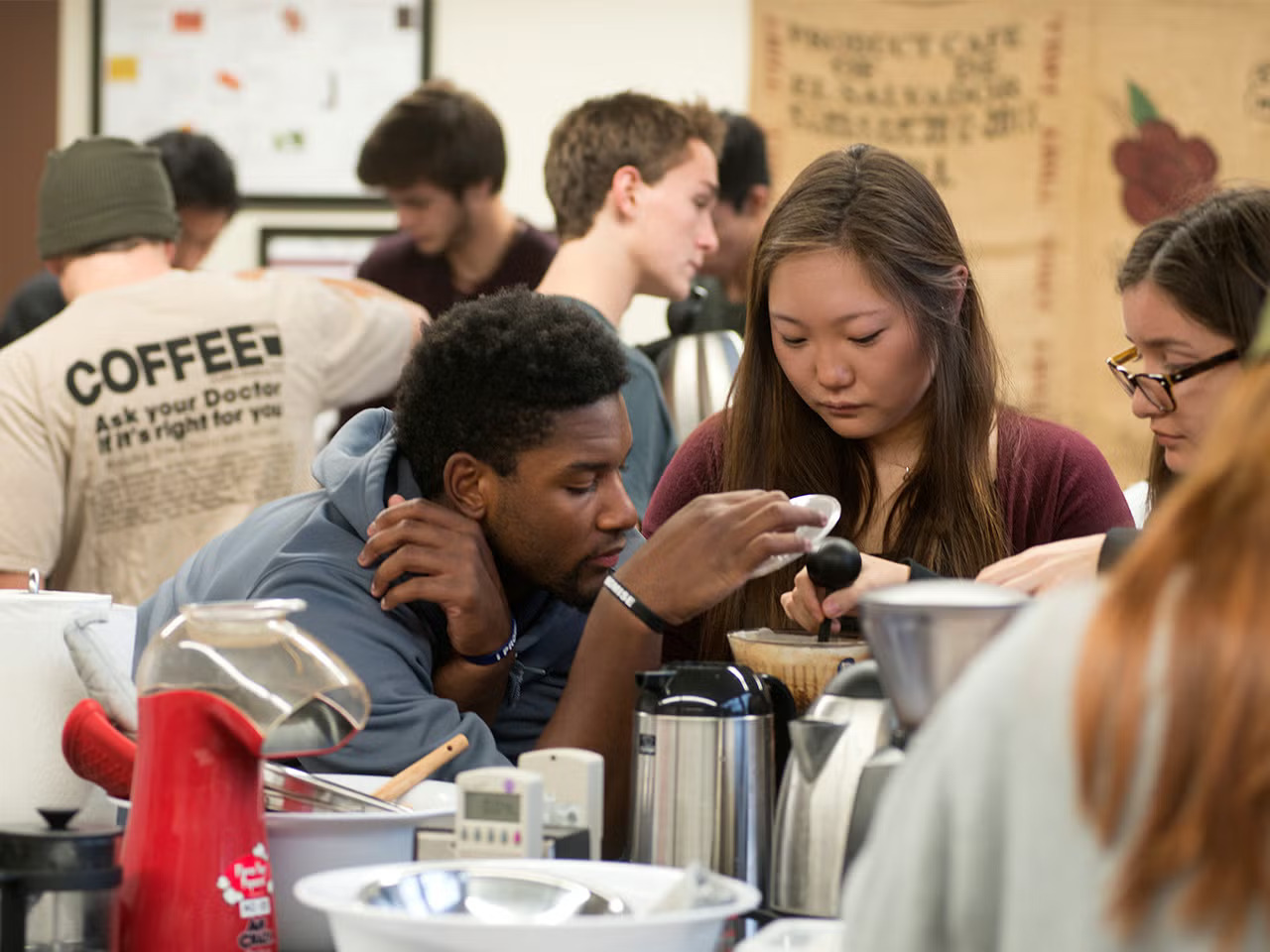 Students work on group projects together; a young Black man delicately holds a piece of equipment over a cone with a coffee filter as a woman looks on and another woman stirs the substance in the coffee filter