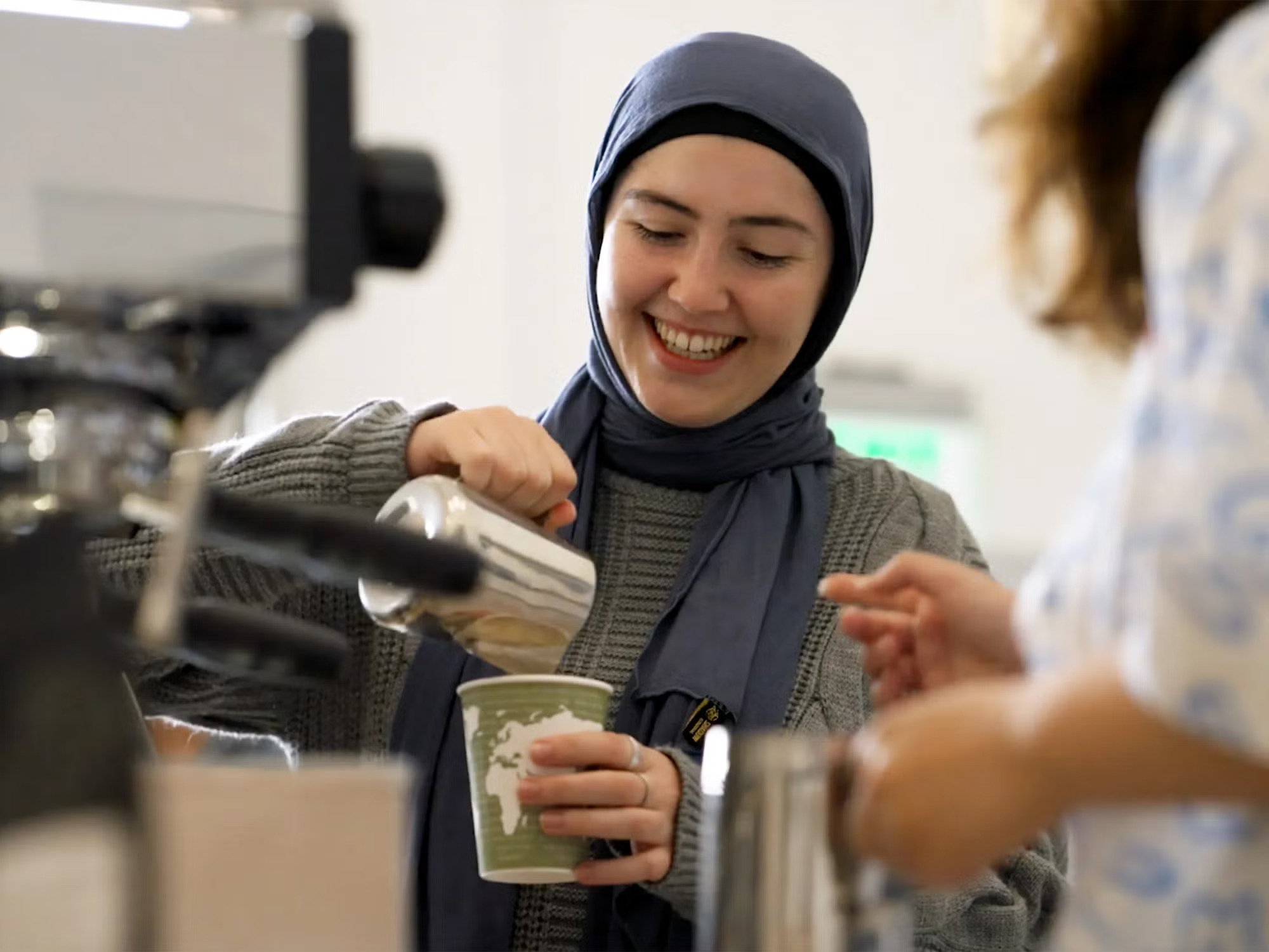 Woman with a headscarf smiles as she pours foam into a paper cup in the coffee lab (other equipment and a person in the foreground)