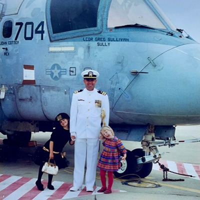 A man in a white Navy uniform holds the hands of two little girls in front of an airplane