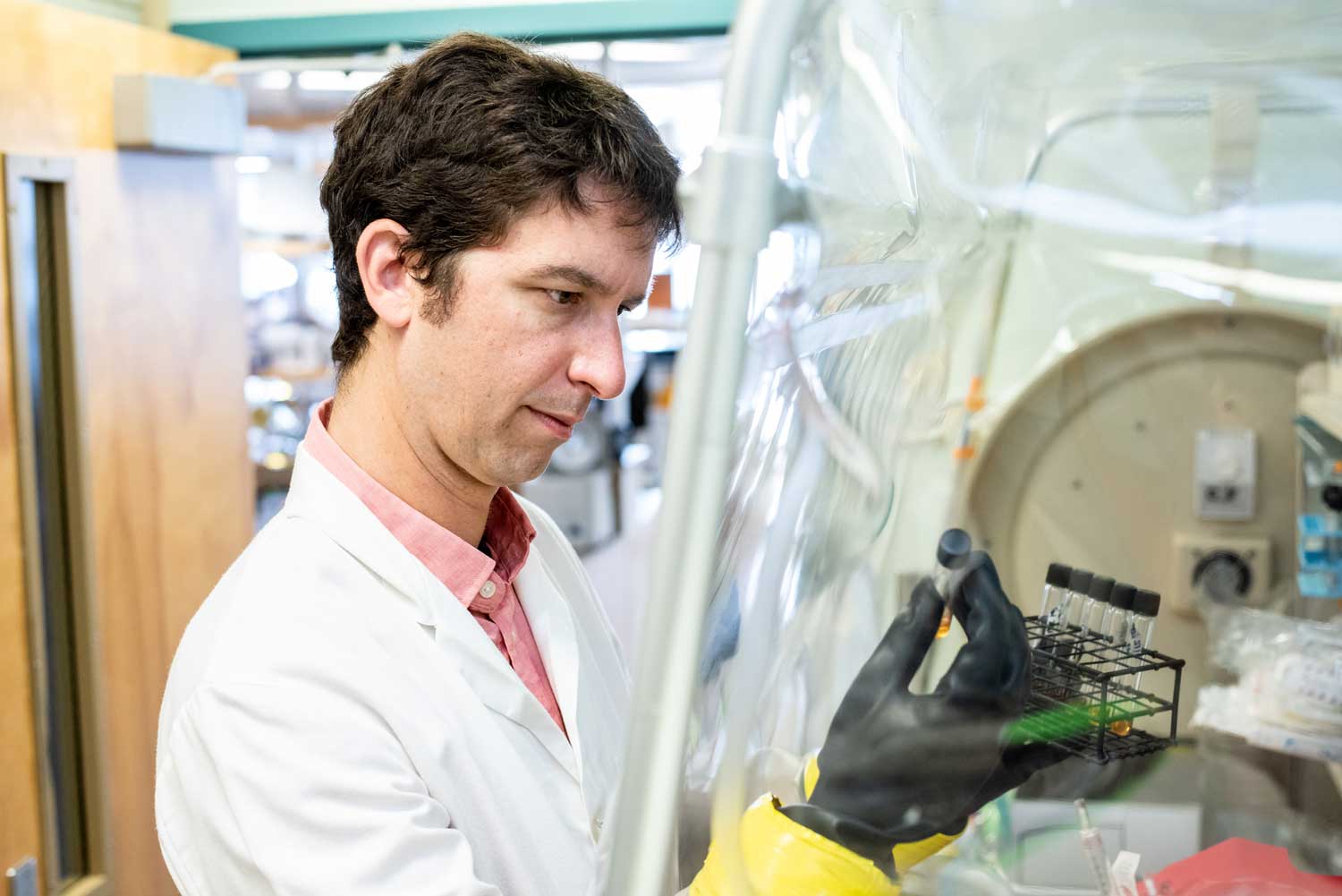 Peter Turnbaugh in a lab, wearing a white coat and black gloves, holding a rack of test tubes, looking closely at one of them