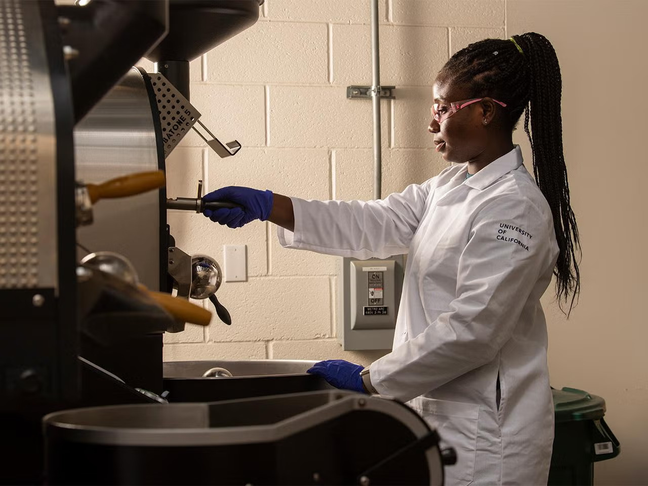 A young Black woman in PPE and a white lab coat with hair pulled back pulls a handle out of a large machine