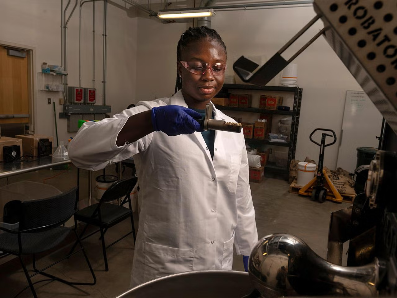 A young Black woman wearing PPE and a white lab coat holds and looks down at a small piece of equipment over a mixer in a lab