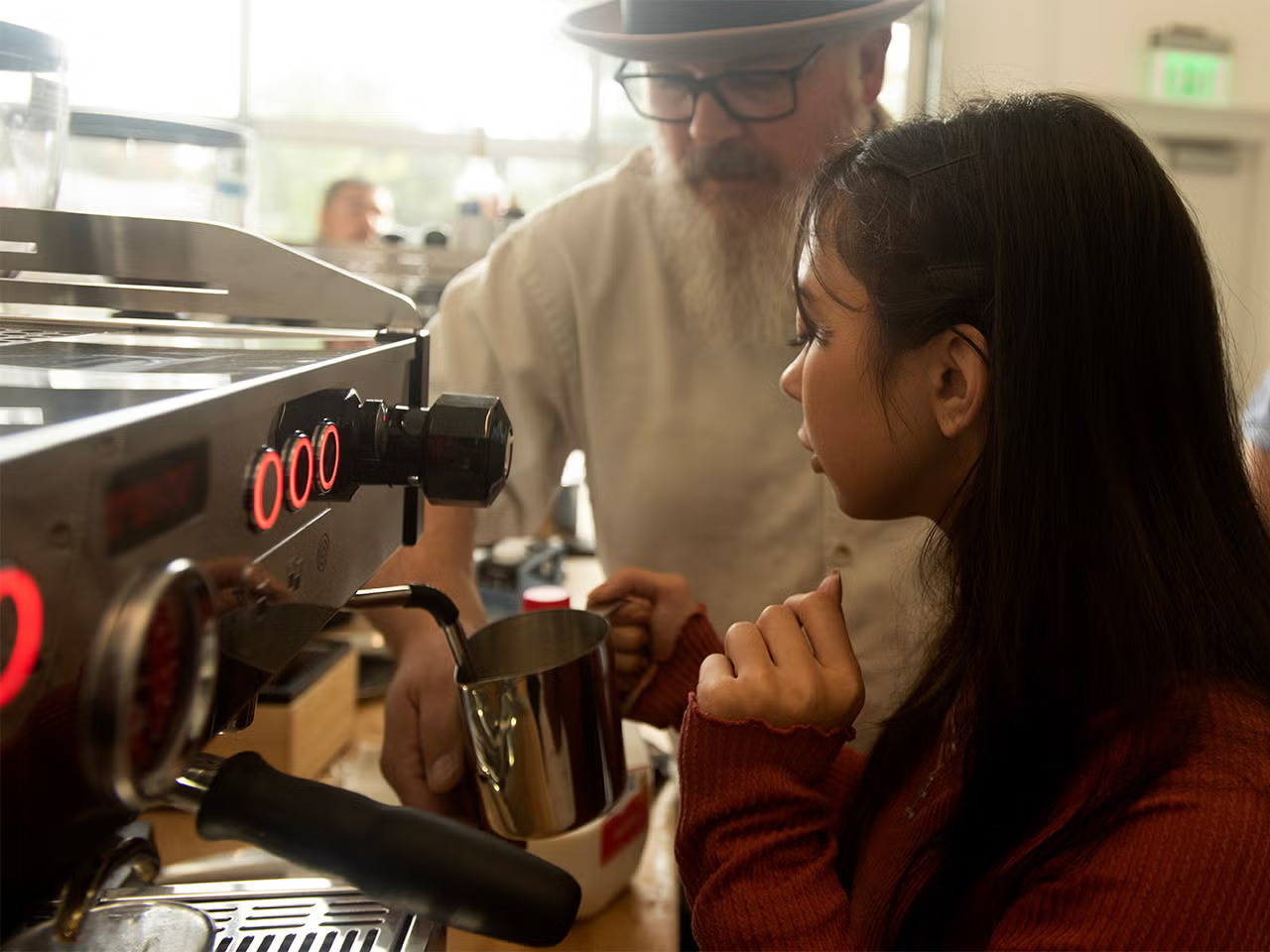 A man with glasses, a fedora and a longish goatee looks at an espresso machine a young woman with long dark hair is operating