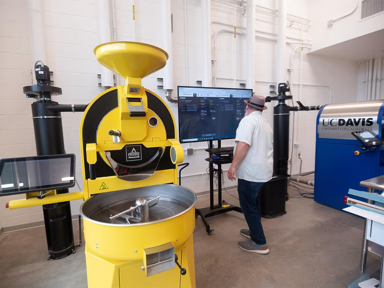 A big yellow coffee roaster with a screen next to it is being operated (by screen) by a man in a Bahama hat and a white shirt 