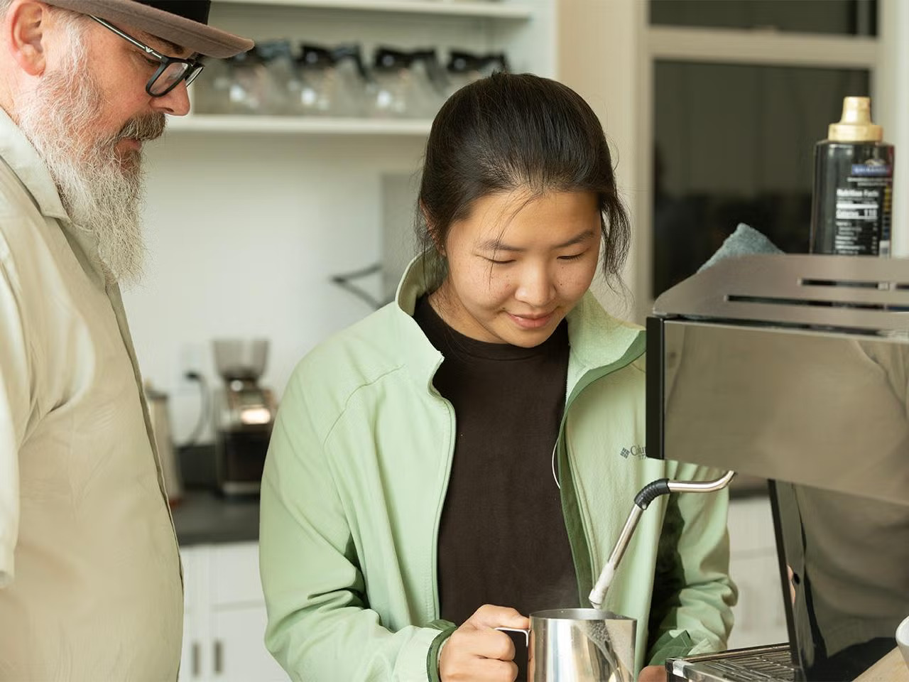 Man with glasses and a Bahama hat and long pointed beard oversees a young woman making coffee from a machine