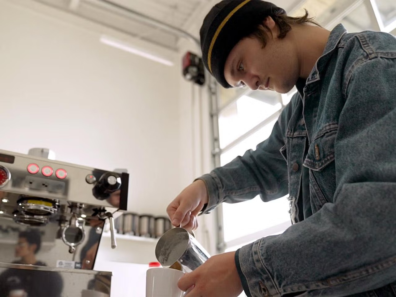 A young man in a jean jacket and beanie pours foam into a coffee cup