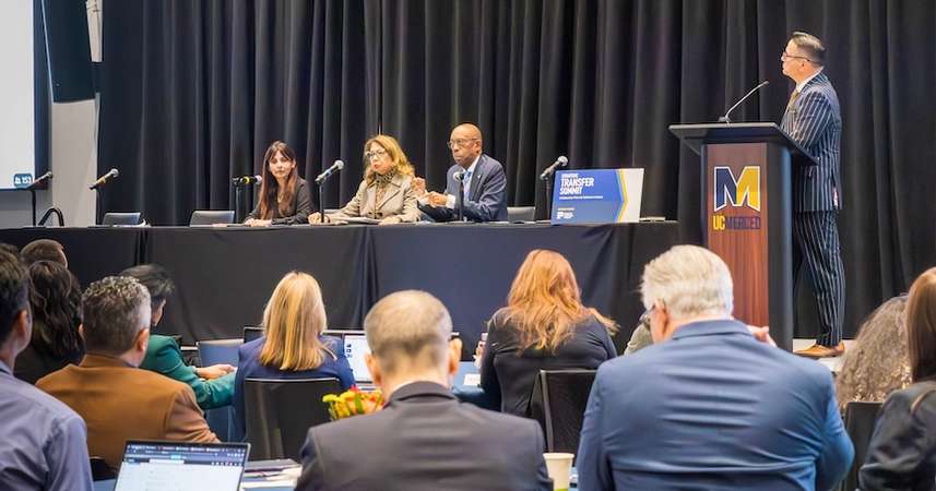 At a table, three people sit in front of microphones, from left: California Community Colleges Chancellor Sonya Christian, California State University Chancellor Mildred Garcia, and UC President Michael Drake; at a lectern, UC Merced Chancellor Juan Sánchez Muñoz stands, looking at the table. Audience is visible a bit in the front of the photo 