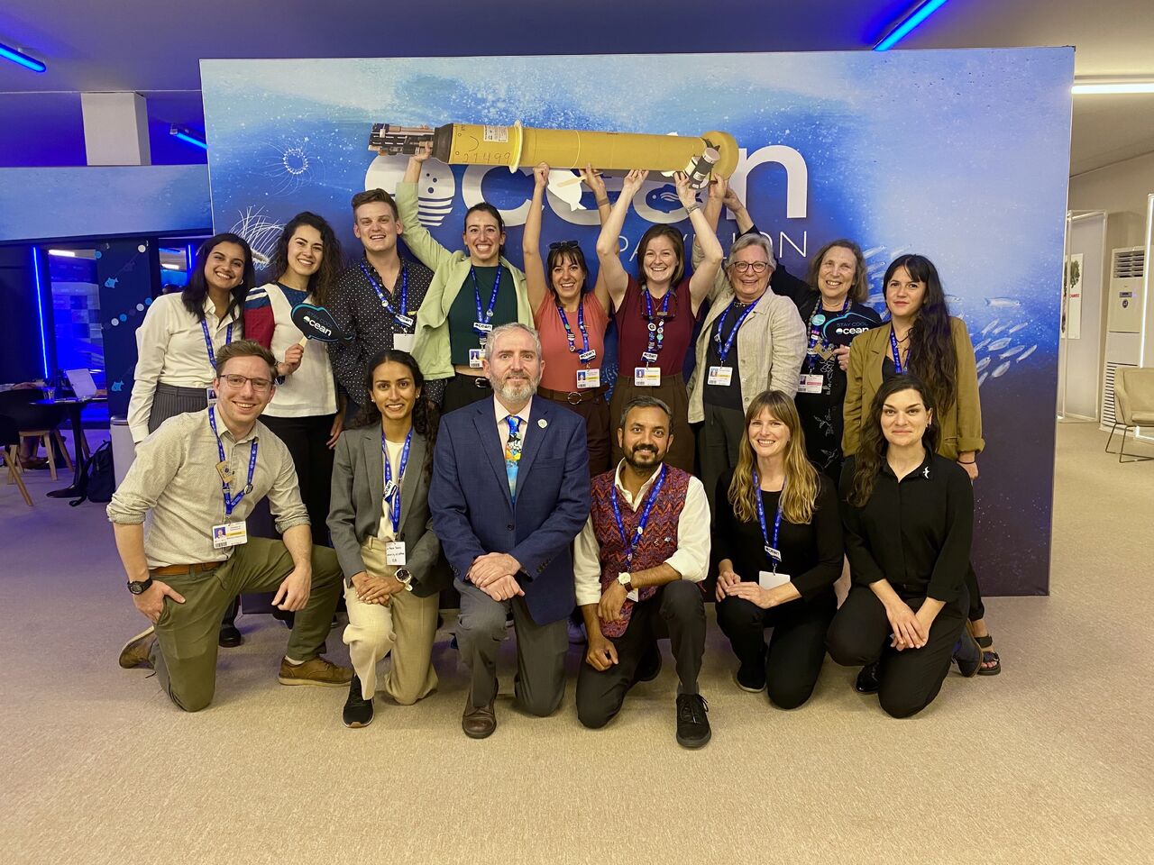 A dozen people in business casual attire pose in two rows in front of a photo backdrop branded "Ocean Pavilion." The back row holds up a yellow cylinder about 4' long above their heads. 