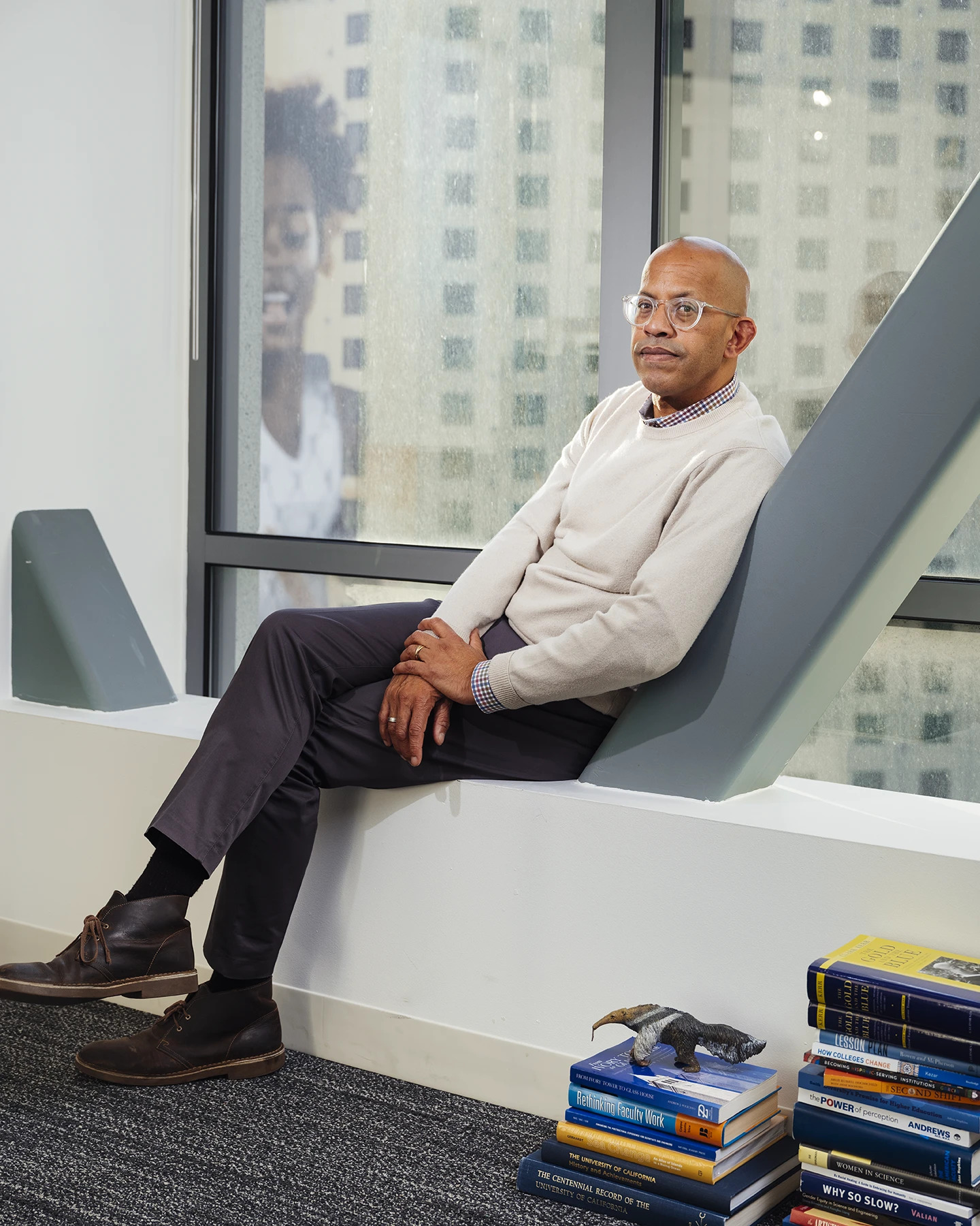 African American man in business casual wear and glasses leaning against a diagonal pillar with a window behind showing a hotel with a mural of a young Black girl (Todd Haynes)