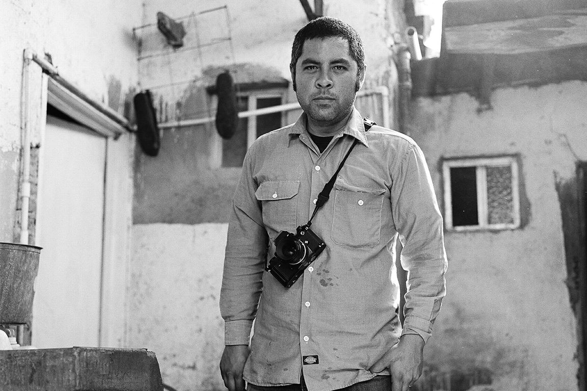 A short-haired man with longish sideburns looks pensively at the camera while in the courtyard of a run down house, camera hanging around his chest