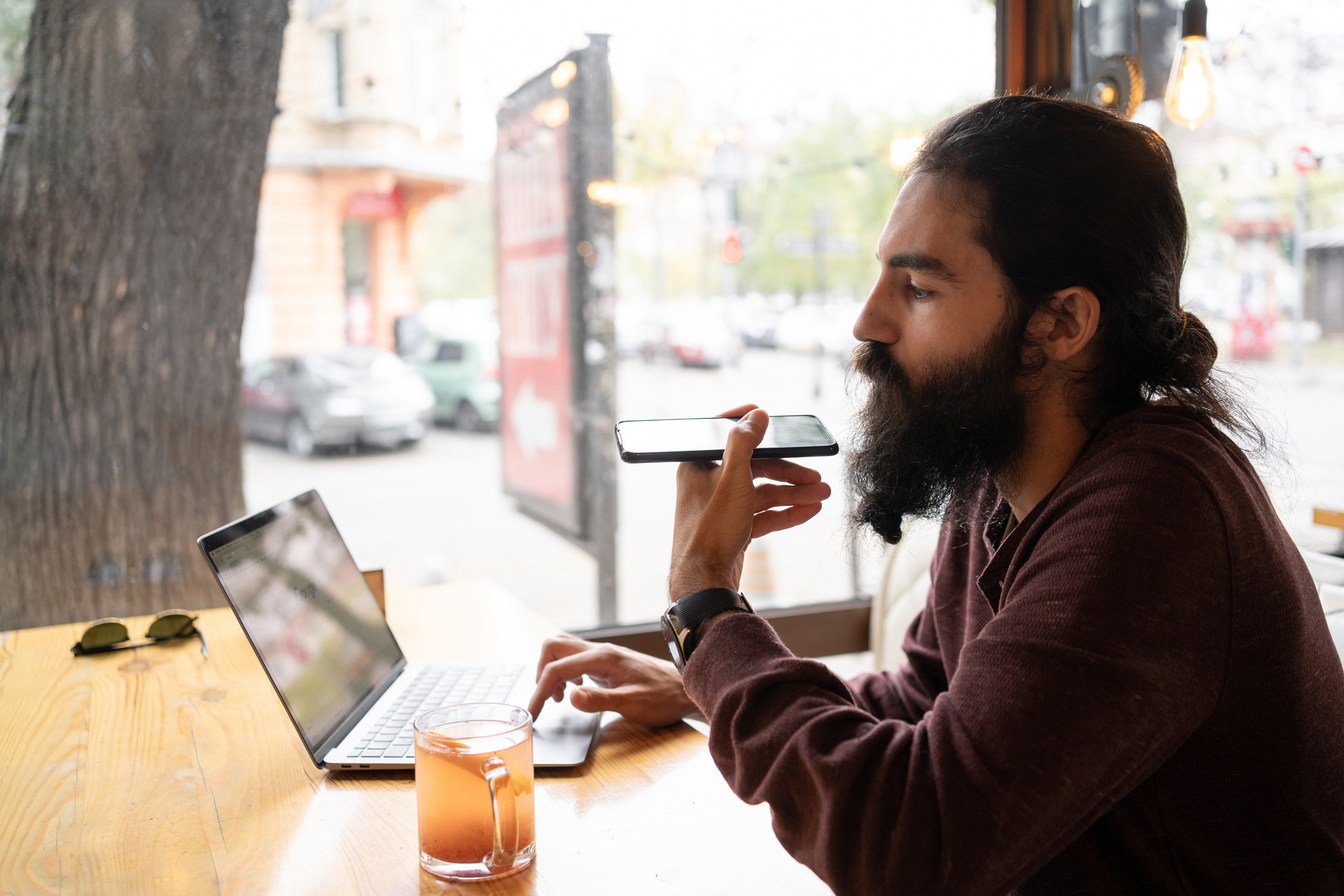 A young bearded man sits at a cafe with a laptop open on the table, speaking into a phone he's holding parallel to the ground