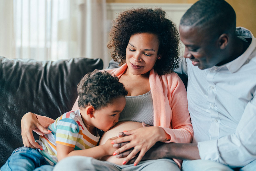 A mom, dad and school-aged kid sit on a couch. The mom is pregnant and the kid is kissing her belly while the dad watches and smiles.