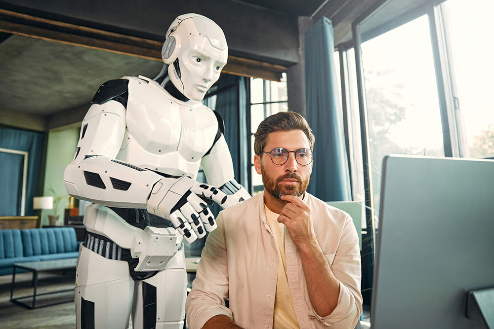 A buff android stands behind a man at a computer, its hand on the man's shoulder. The android is pointing toward the screen and the man has a thoughtful expression, as if listening.