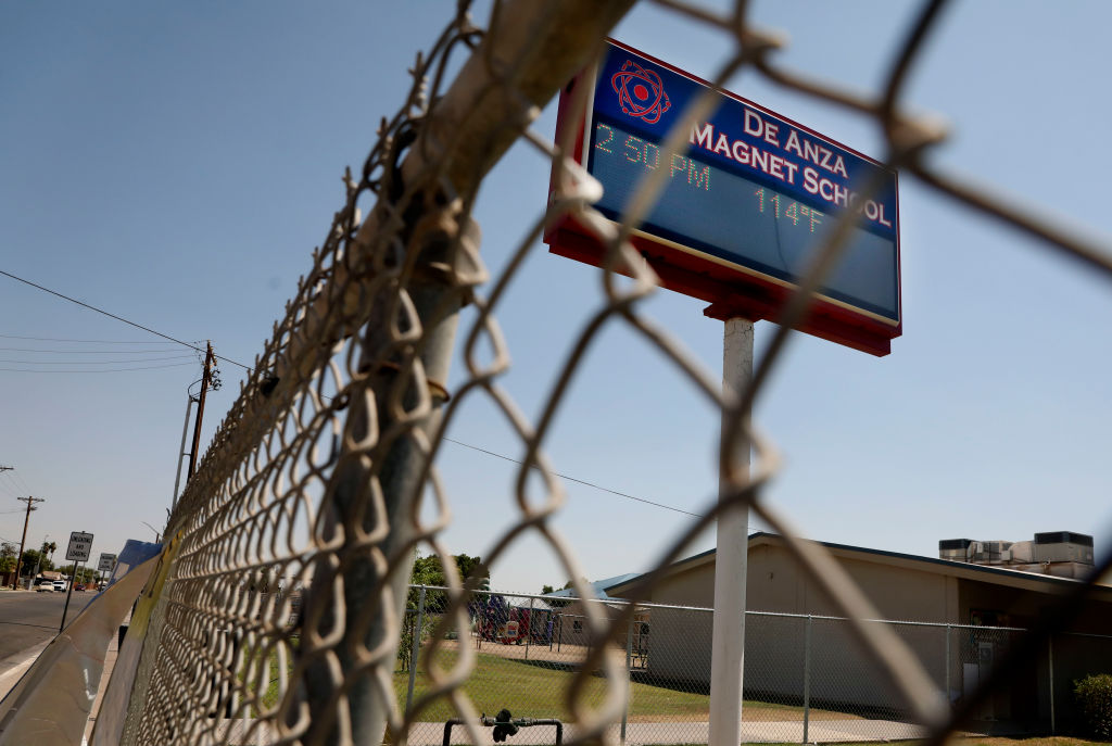 A temperature of 114 degrees F is displayed on a digital sign outside of De Anza Magnet School, photographed through a chain link fence.